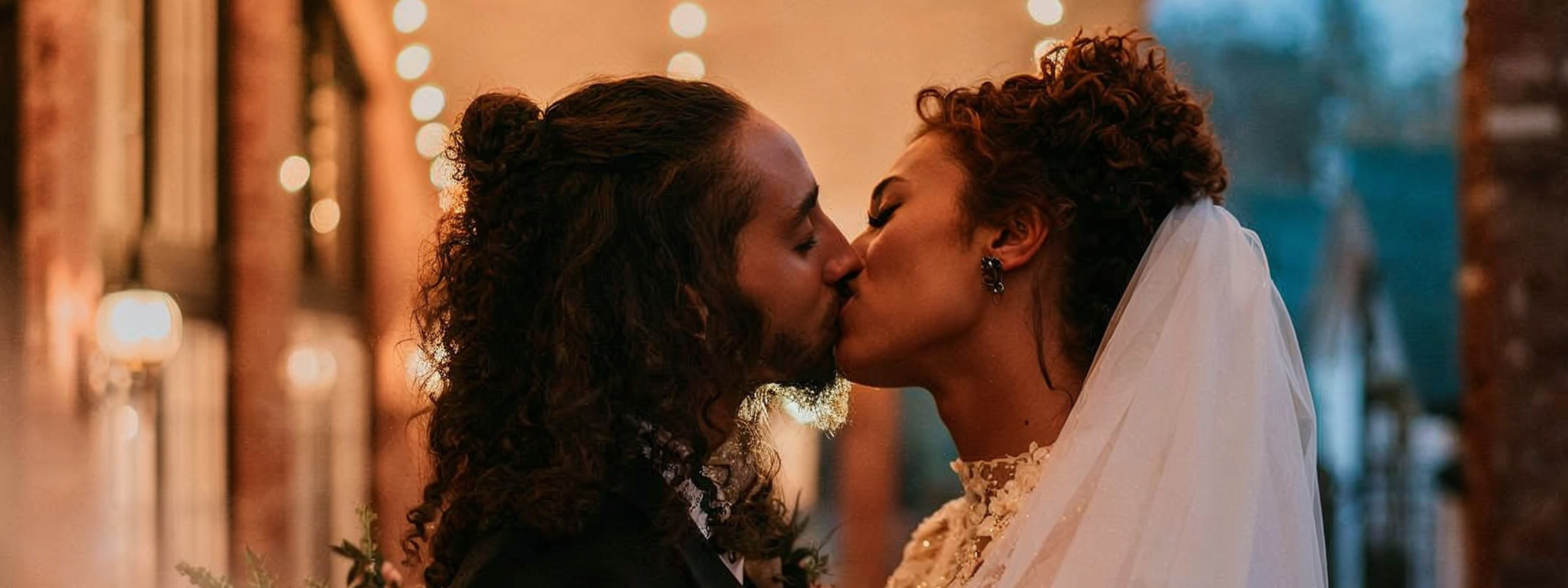 A newlywed couple shares a romantic kiss. The bride wears Amity Alexandra's Thorned Heart Earrings, complementing her elegant lace wedding gown and flowing veil.