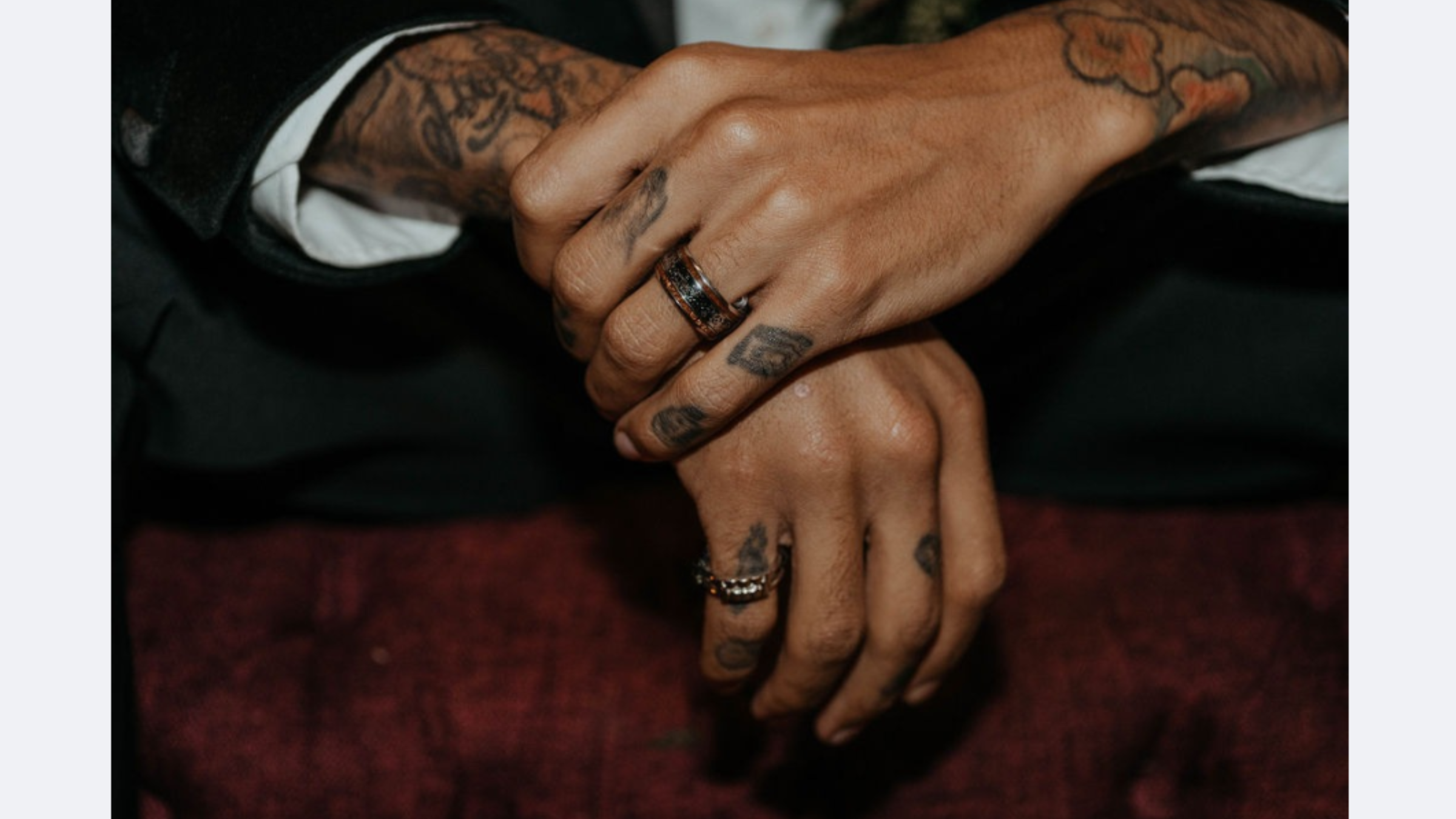 Close-up of tattooed hands wearing custom men's wedding bands, including a black and wood inlay ring and a textured silver band.